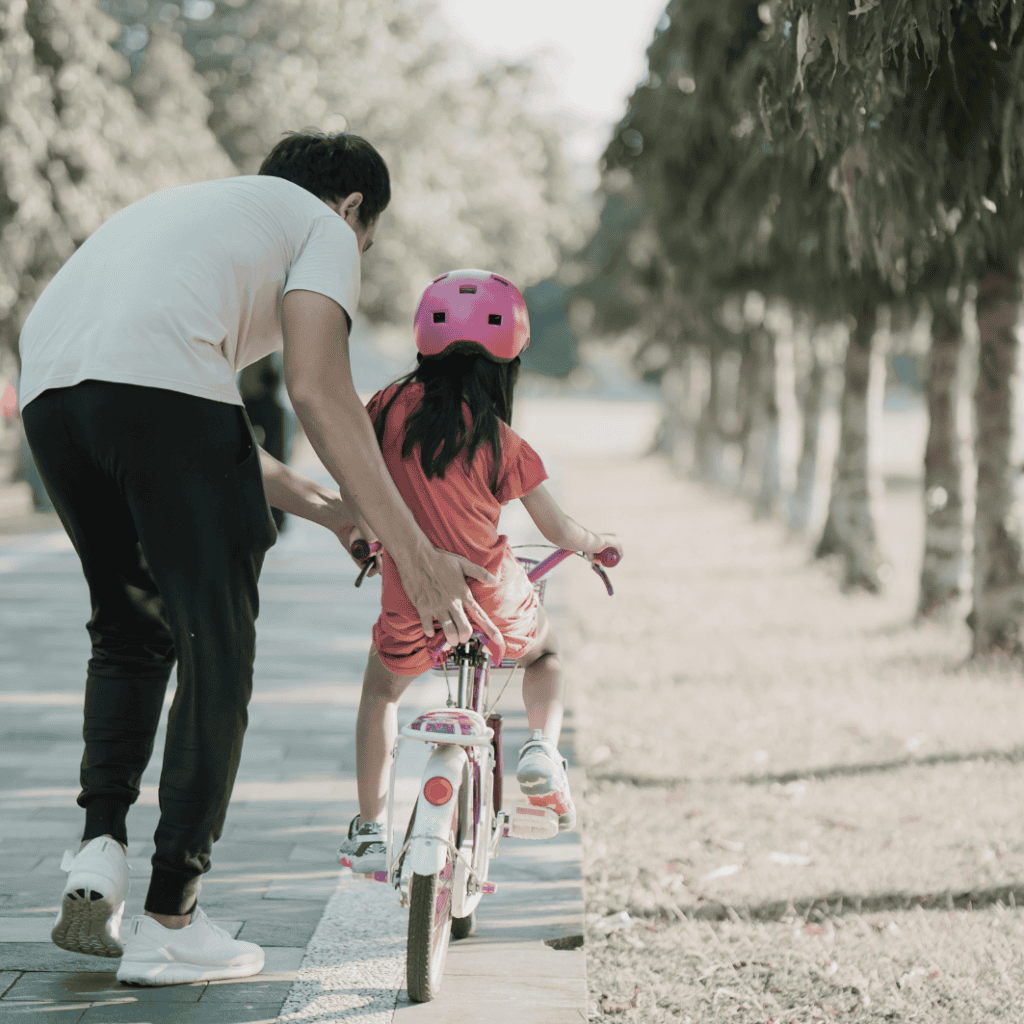Father helping daughter ride bike on park path with trees.