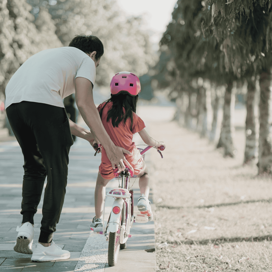 Father helping daughter ride bike on park path with trees.
